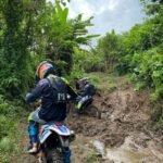 A dirt biker riding uphill on a rocky trail surrounded by lush green vegetation.